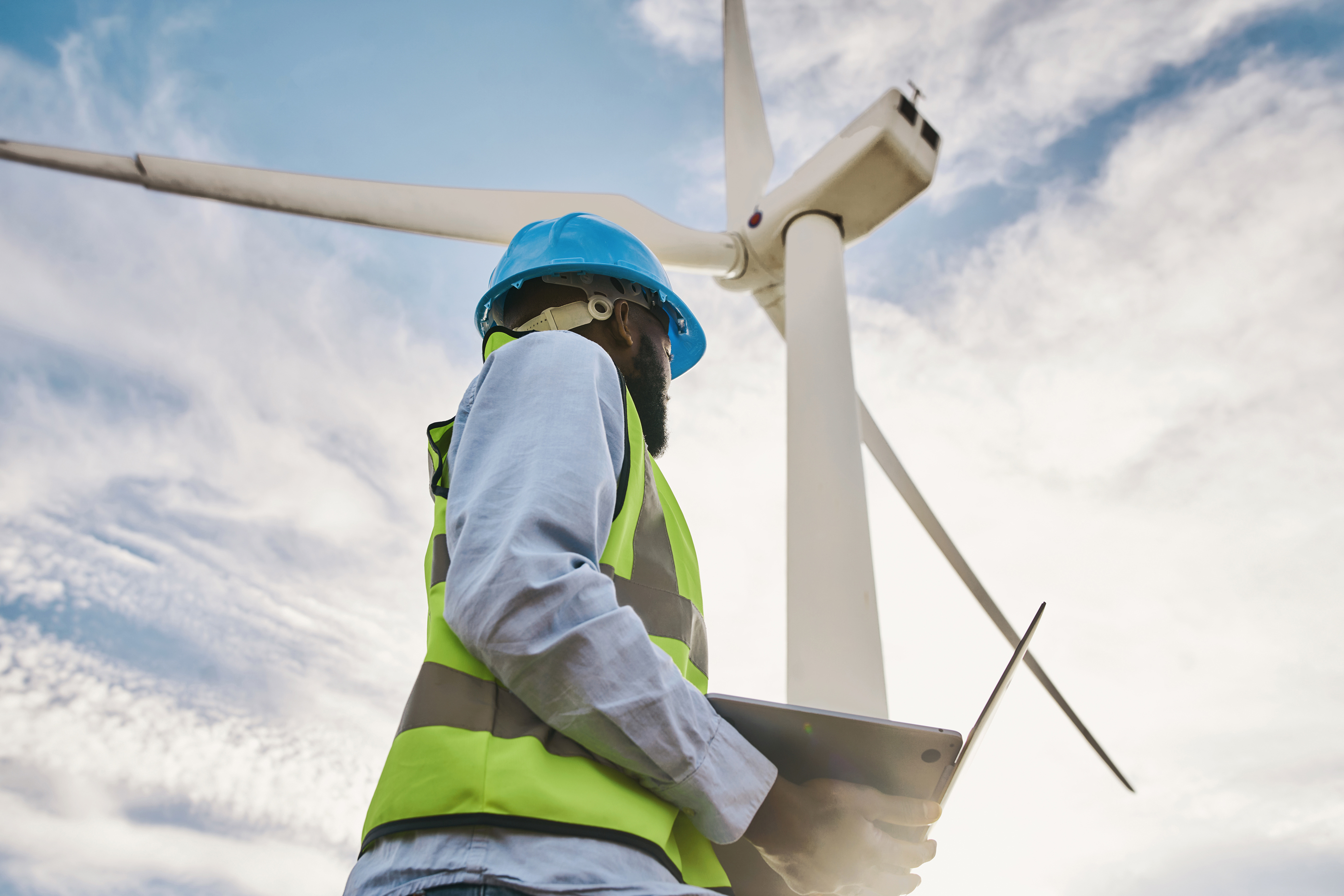 Worker in bright blue hard hat standing beneath a wind turbine.