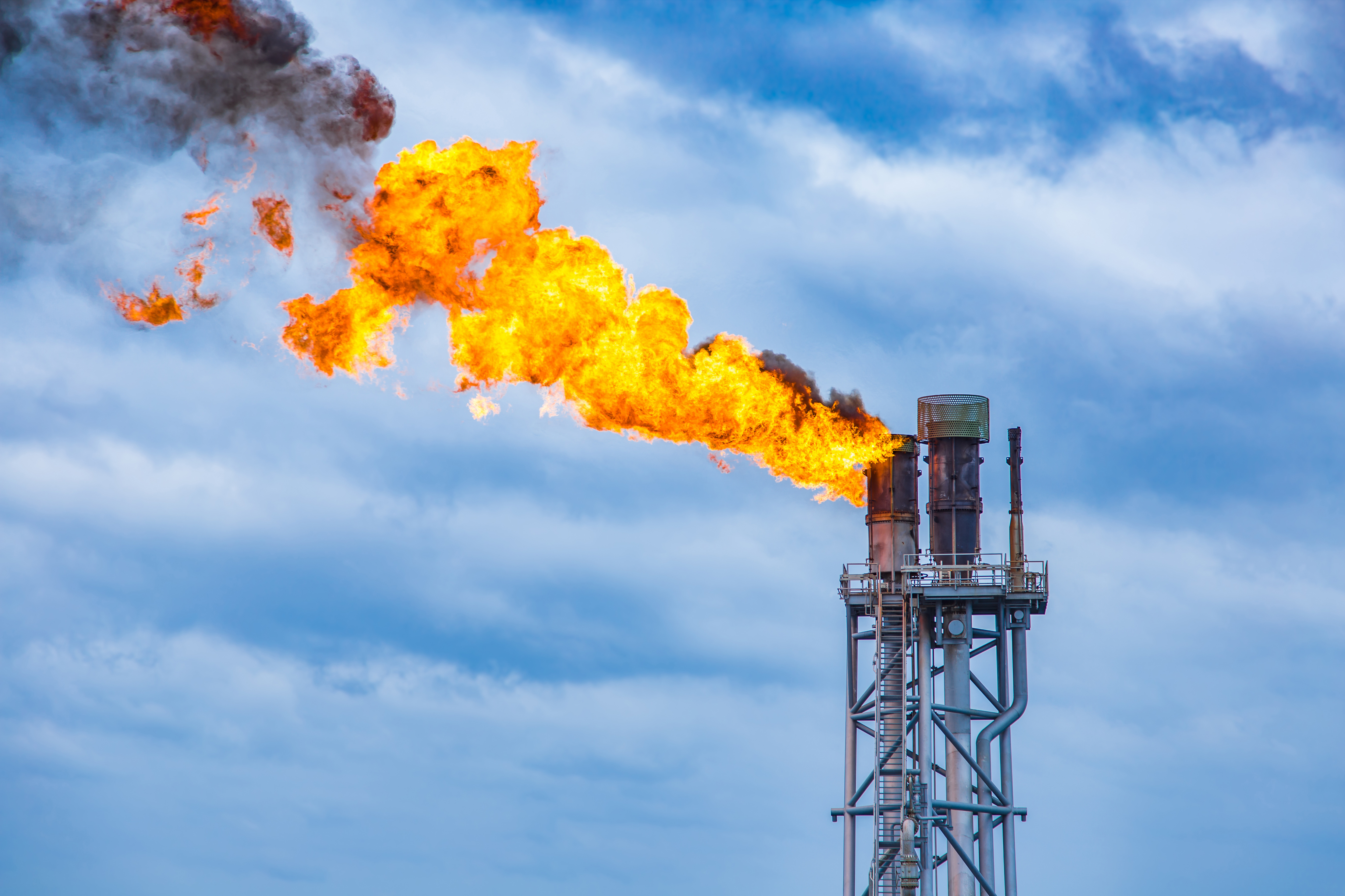 A tall industrial flare stack burns a large, bright orange flame against a cloudy blue sky, with dark smoke drifting from the fire.