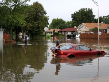A man sitting on top of car in the middle of flooded area.